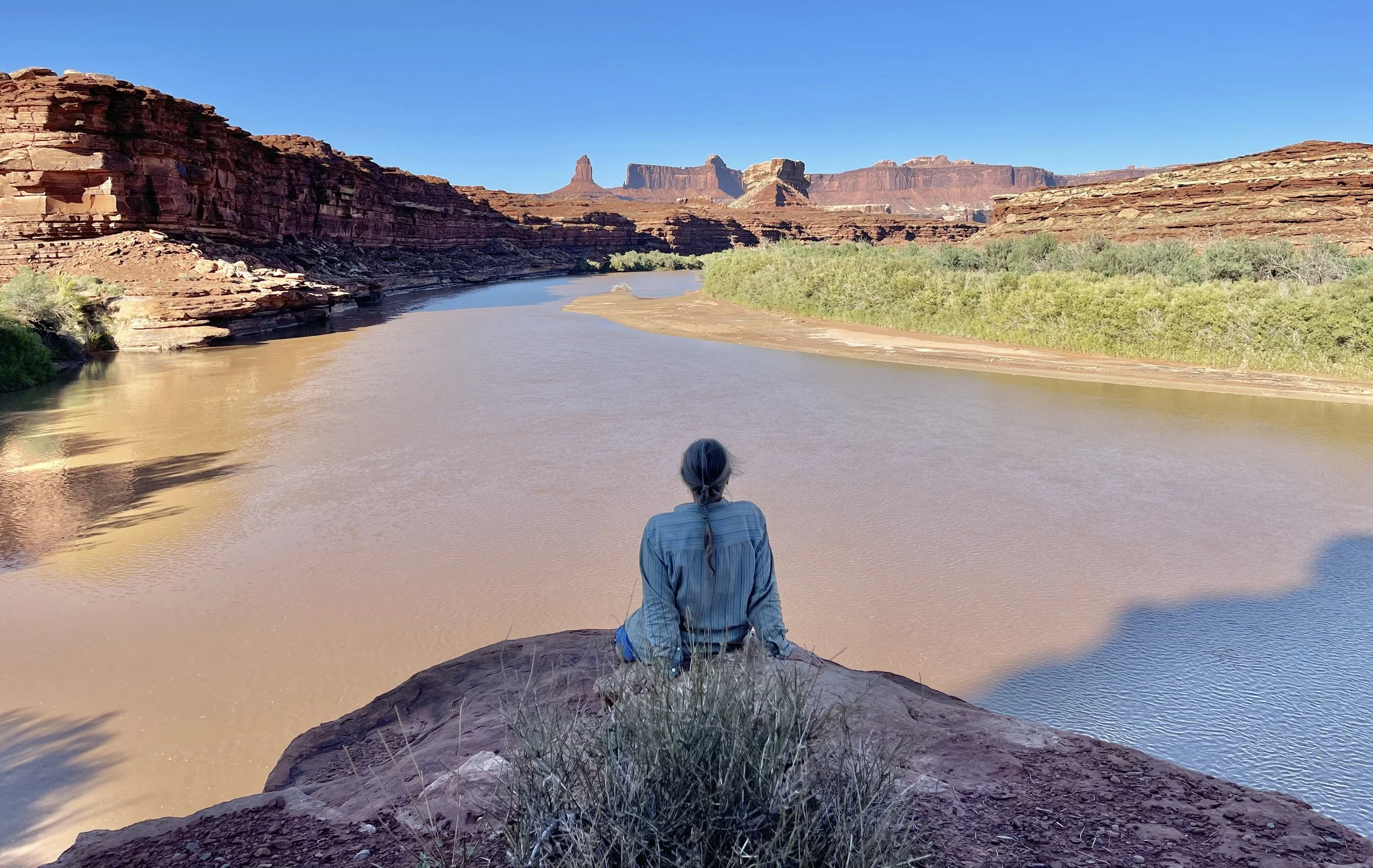 A woman sitting on a rock by a river in a desert canyon, overlooking towering red rock formations under a clear blue sky.