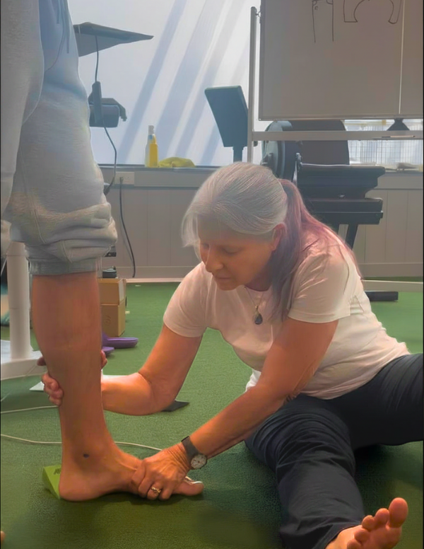 A woman with gray hair and a white shirt is performing physical therapy or a stretching exercise on a person's lower leg and foot, who is standing with their foot on a green floor in a fitness or therapy room.
