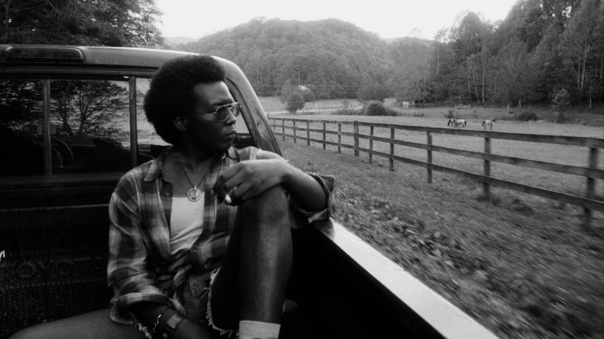 A black man in a flannel and denim shorts smokes a cigarette while watching horse pastures from a moving truckbed.