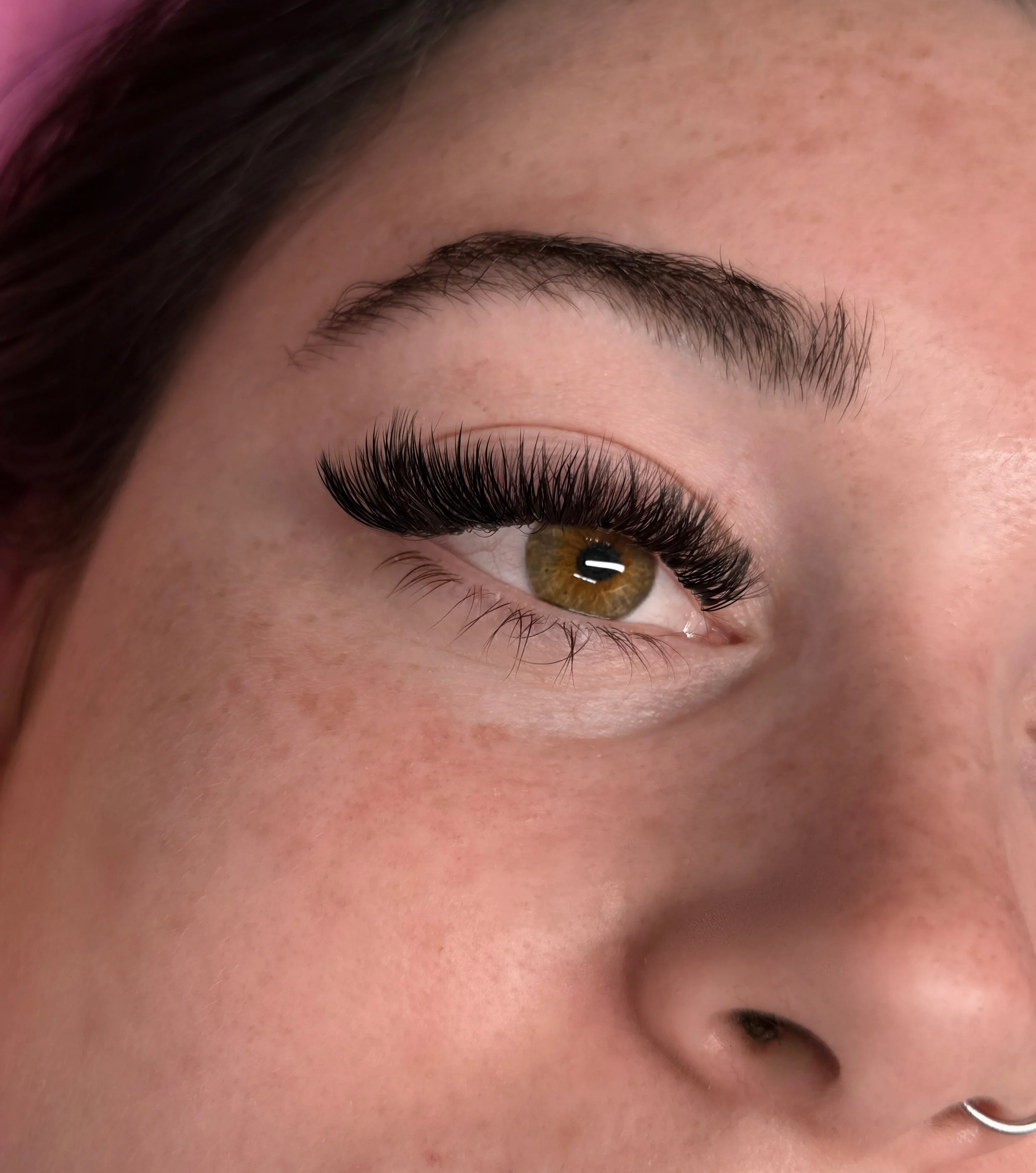 Close-up of a person with long, thick false eyelashes, showing their eye, eyebrow, and part of their nose and cheek.