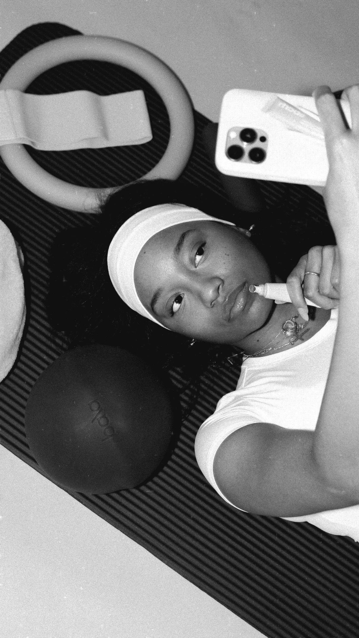 Black and white photograph of a young woman lying on the floor in workout attire, holding a phone and taking a selfie. She wears a white headband and has fitness equipment around her, including a large ring, a ball, and a foam roller, on a striped workout mat.