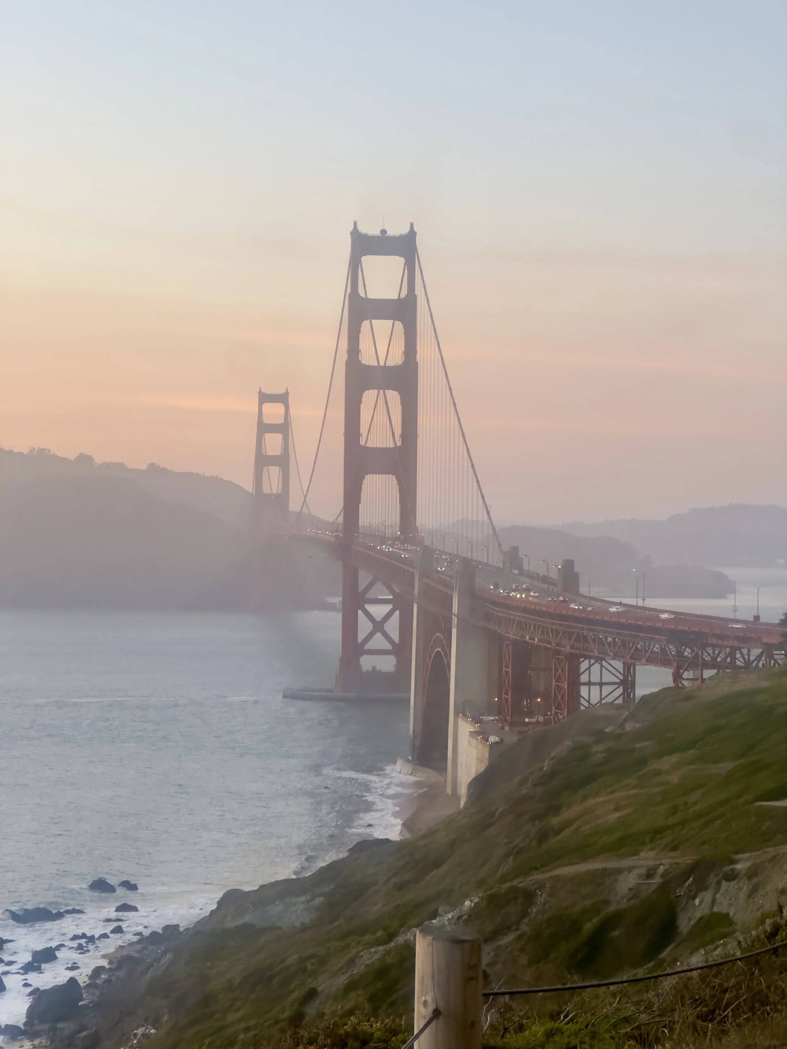 The Golden Gate bridge extending over a body of water with hills and coastline in the background at sunset or sunrise.
