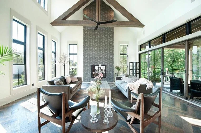 Bright modern living room with large windows, a gray sofa, black chairs, and a glass door leading to an outdoor patio.