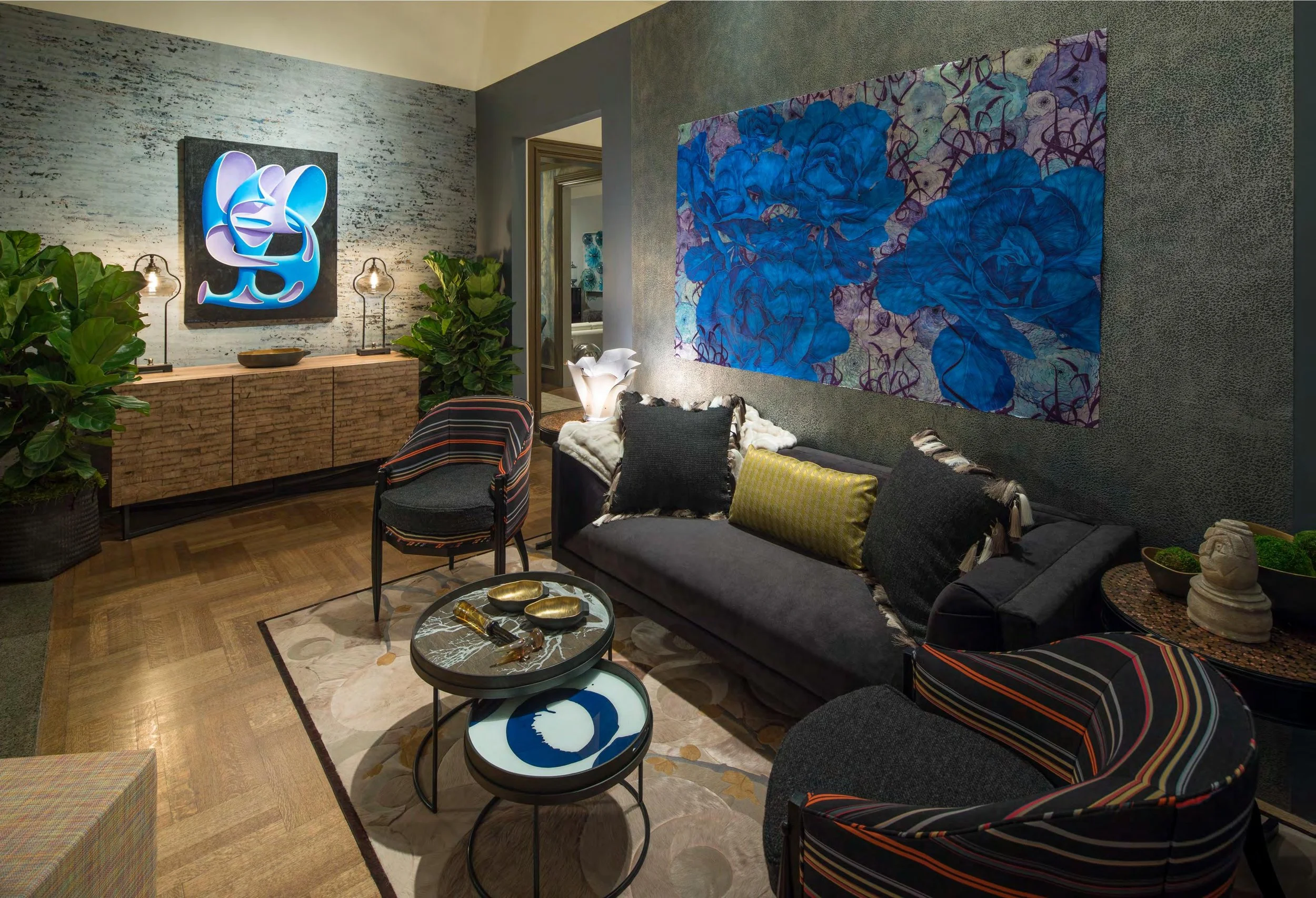 A modern living room with a black sofa adorned with colorful cushions, a wooden sideboard with lamps, large green plants, and abstract artwork on the walls, including a blue floral painting.