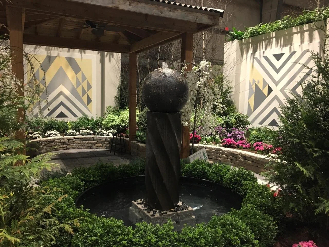 A small garden with a fountain featuring a black spherical top and a twisted black stone pillar, surrounded by lush green bushes, blooming flowers, and a covered seating area with patterned walls in the background.