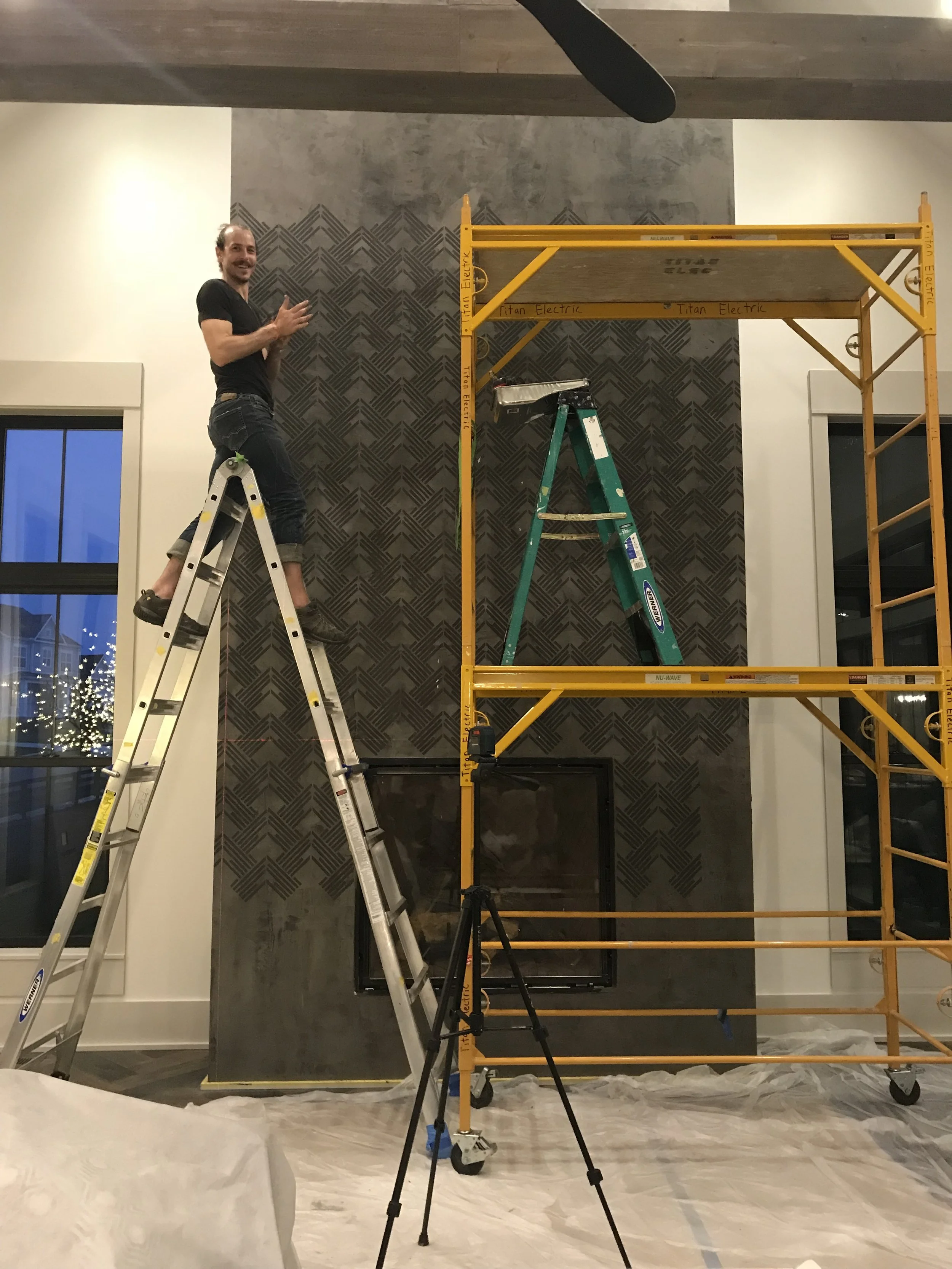 Jesse Getic standing on a ladder in front of a wall with textured gray chevron design, using scaffolding to reach higher. A tripod is set up on the floor, and there is a window showing a festive outdoor scene with decorative lights.