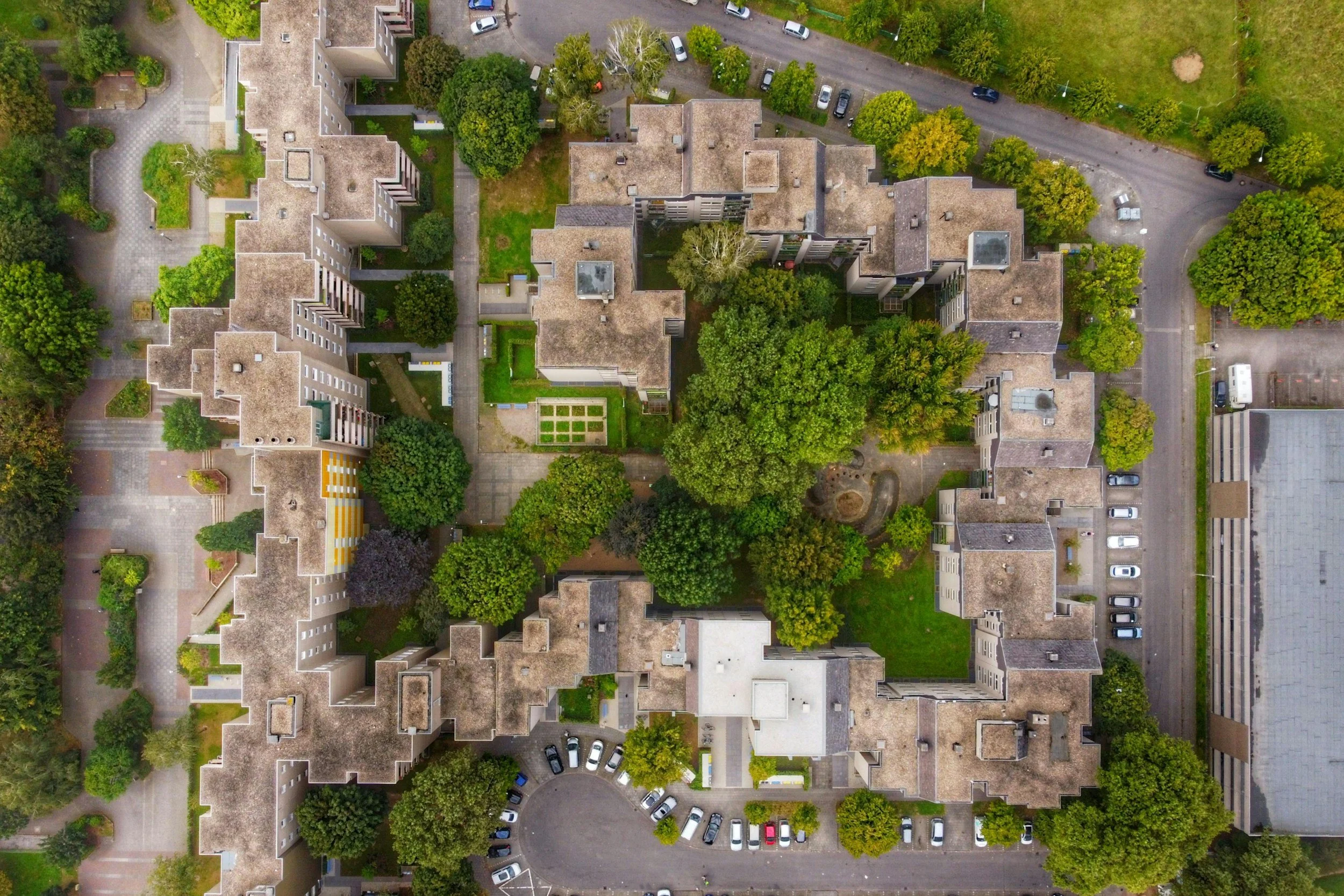 An aerial view of residential buildings with rooftops, green trees, driveways, and parked cars.