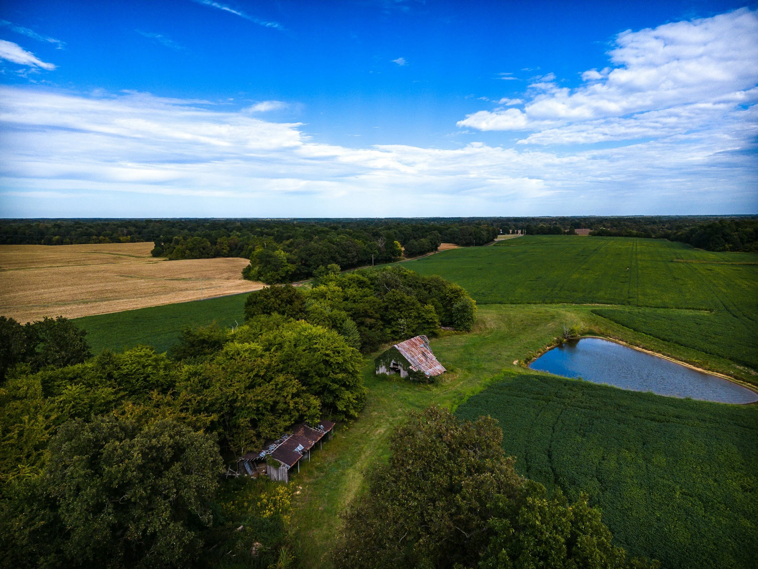 A vast rural landscape with fields, trees, a pond, and an old barn, under a partly cloudy blue sky.