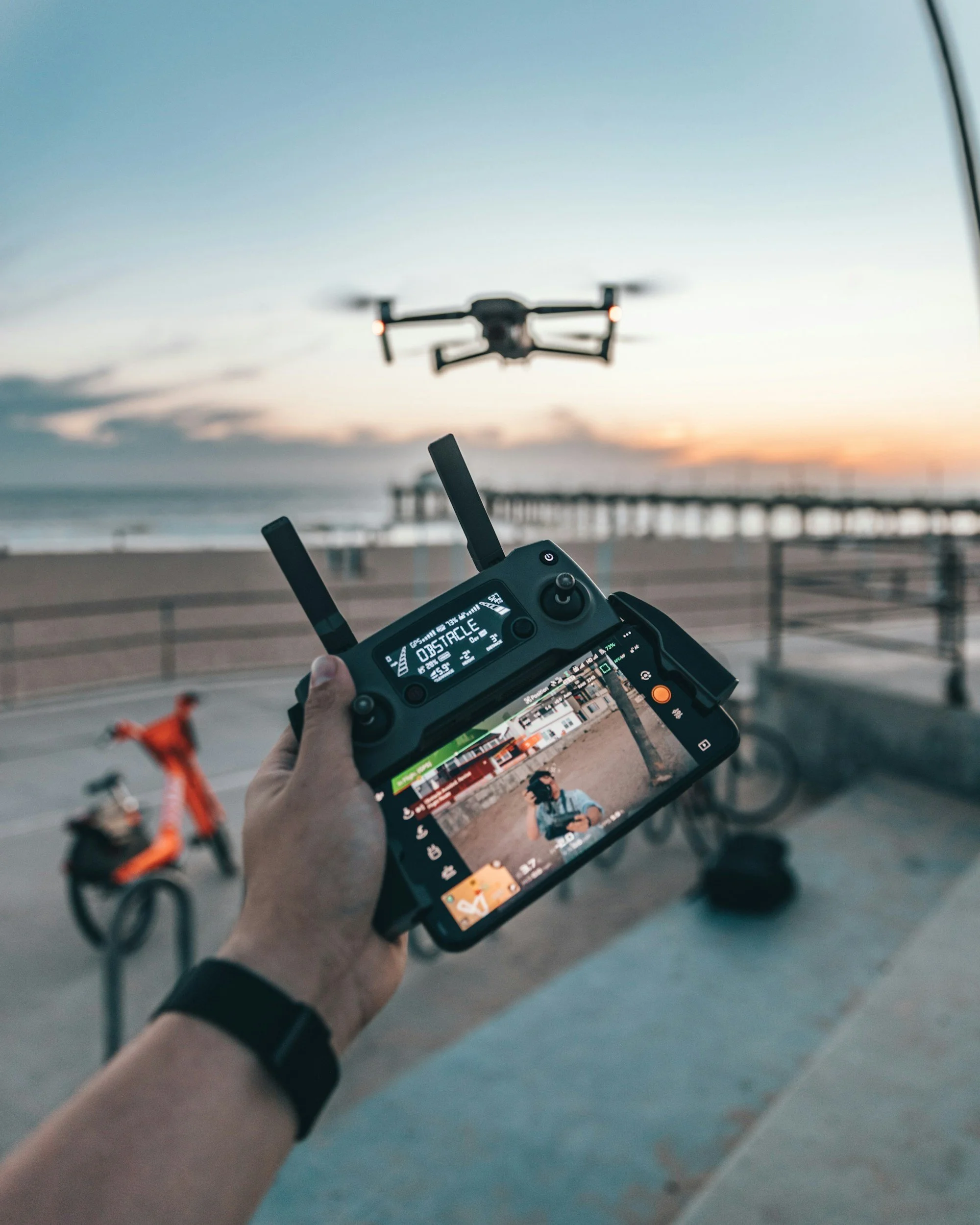 Person holding a drone transmitter with a drone flying in the sky at sunset in the background, a person on a bicycle in the distance.