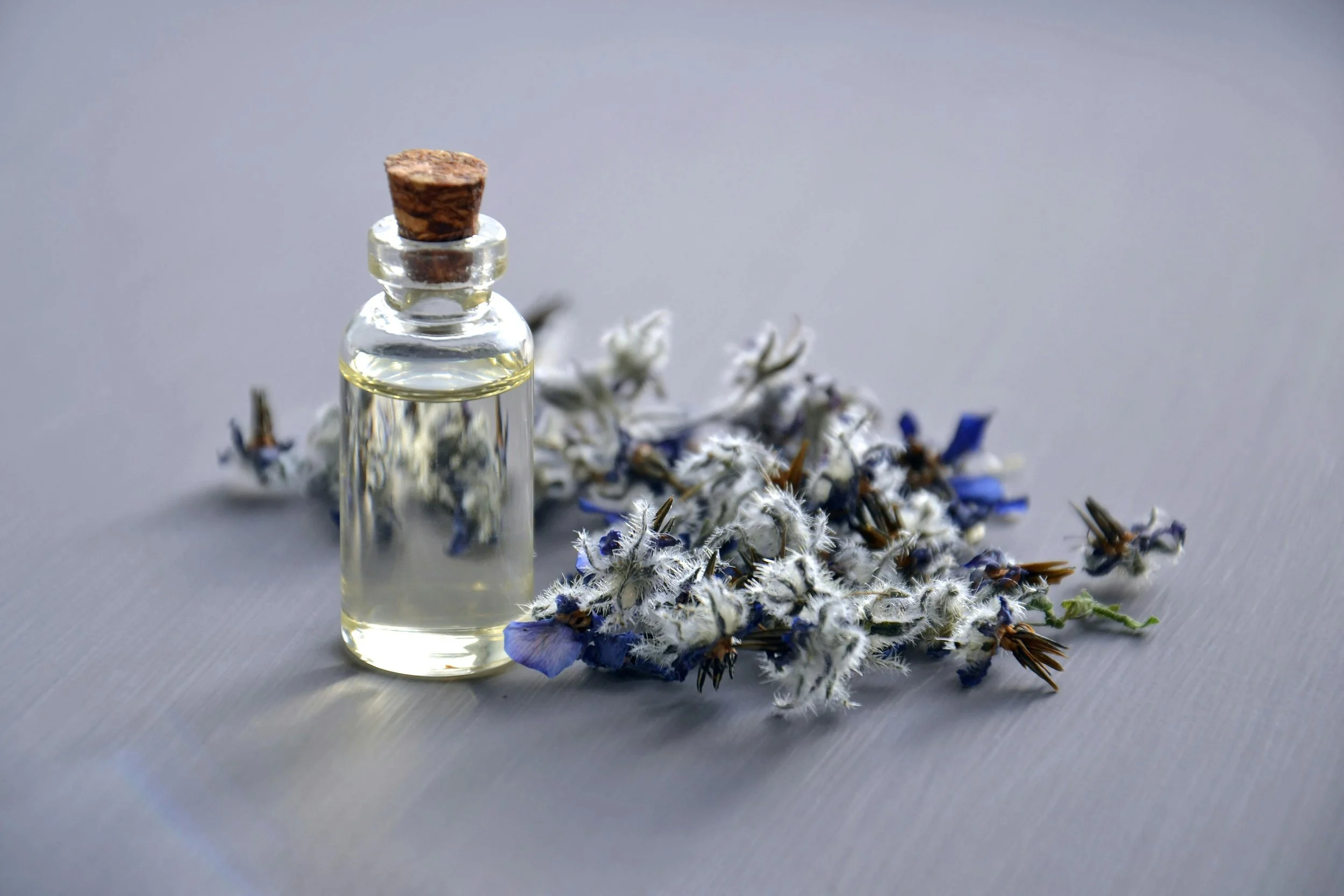 A small glass bottle containing a clear liquid, sealed with a cork, placed on a surface with dried blue and white flowers.