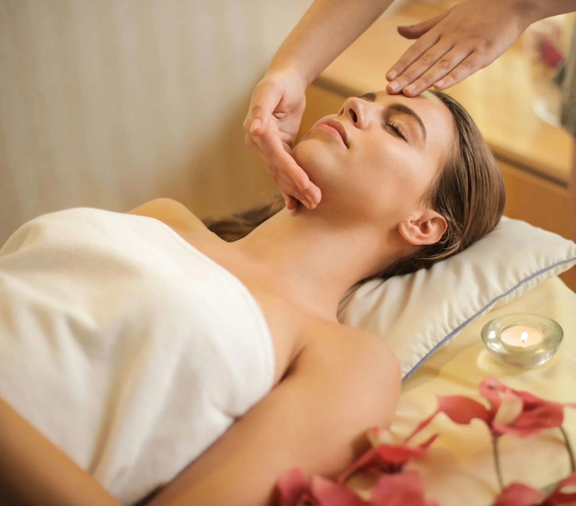 A woman lying on a massage table receiving a facial massage, with pink flowers and a lit candle nearby in a spa room.