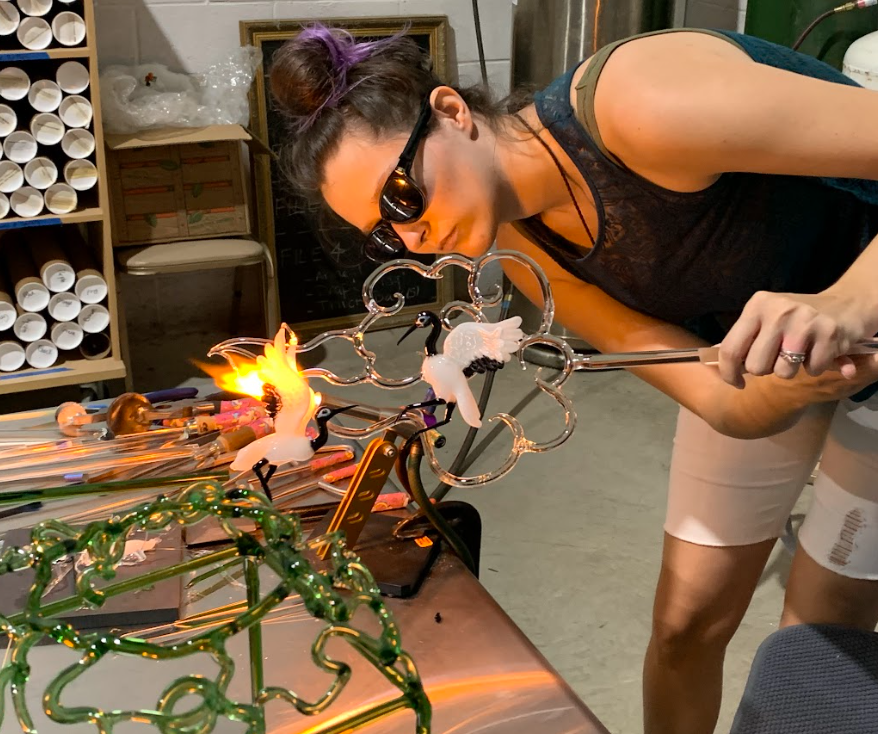 A woman with dark hair in a bun, wearing sunglasses, black sleeveless top, and beige shorts, is blowing glass into a decorative glass object shaped like a flower with swan figures, in a glass studio with various glass art tools and supplies on the table.