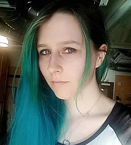 Close-up of a young woman with long hair that's dark at the top and green at the ends, in an indoor setting.