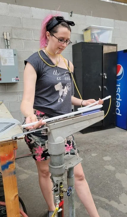 Young woman with pink hair in a black headband, glass, and casual clothing uses a machine in a workshop or industrial setting.