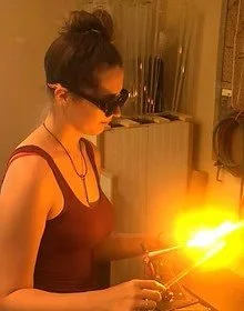 A woman wearing safety glasses holds a welding torch with bright sparks in a workshop.