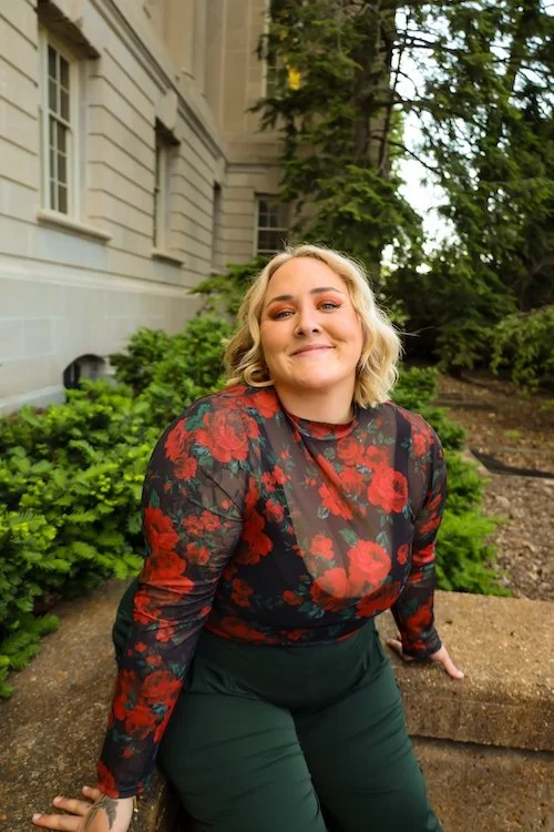 A woman with blonde hair smiling, wearing a black sheer blouse with red floral pattern and green pants, sitting outdoors on a stone ledge with greenery and trees in the background.