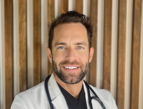 Smiling male doctor with stethoscope, wearing white coat, standing in front of wooden panel background.