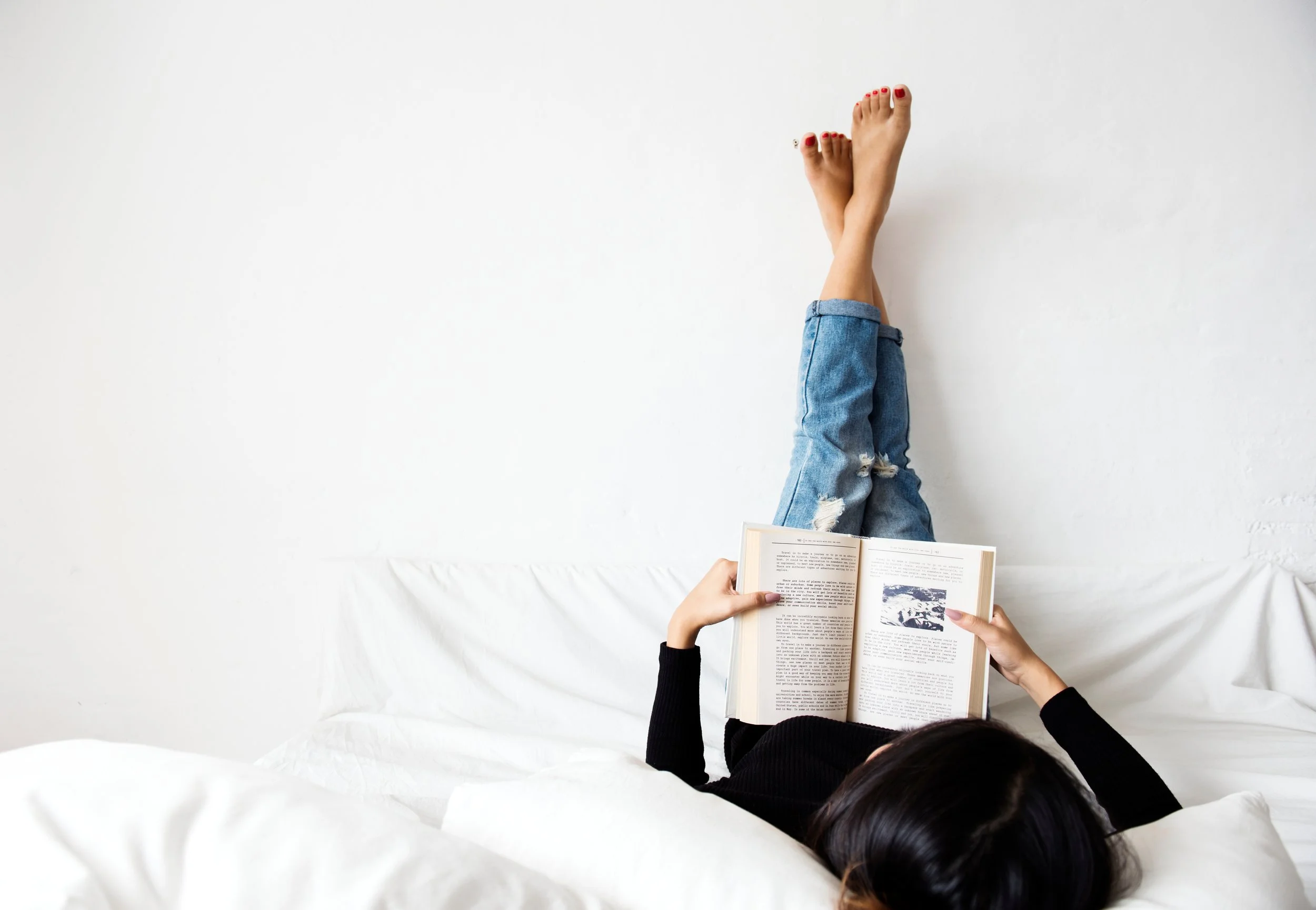Person lying on a bed with legs up against the wall, reading a book, wearing blue ripped jeans and a black sweater, with red nail polish.