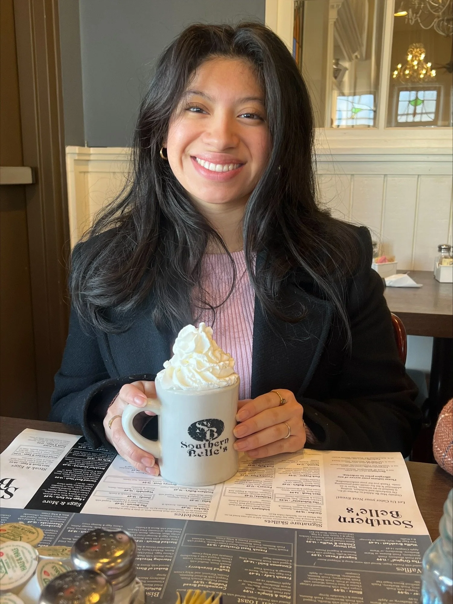 A woman smiling while holding a coffee mug topped with whipped cream. She is seated at a table in a restaurant or cafe, with a menu and condiments on the table.