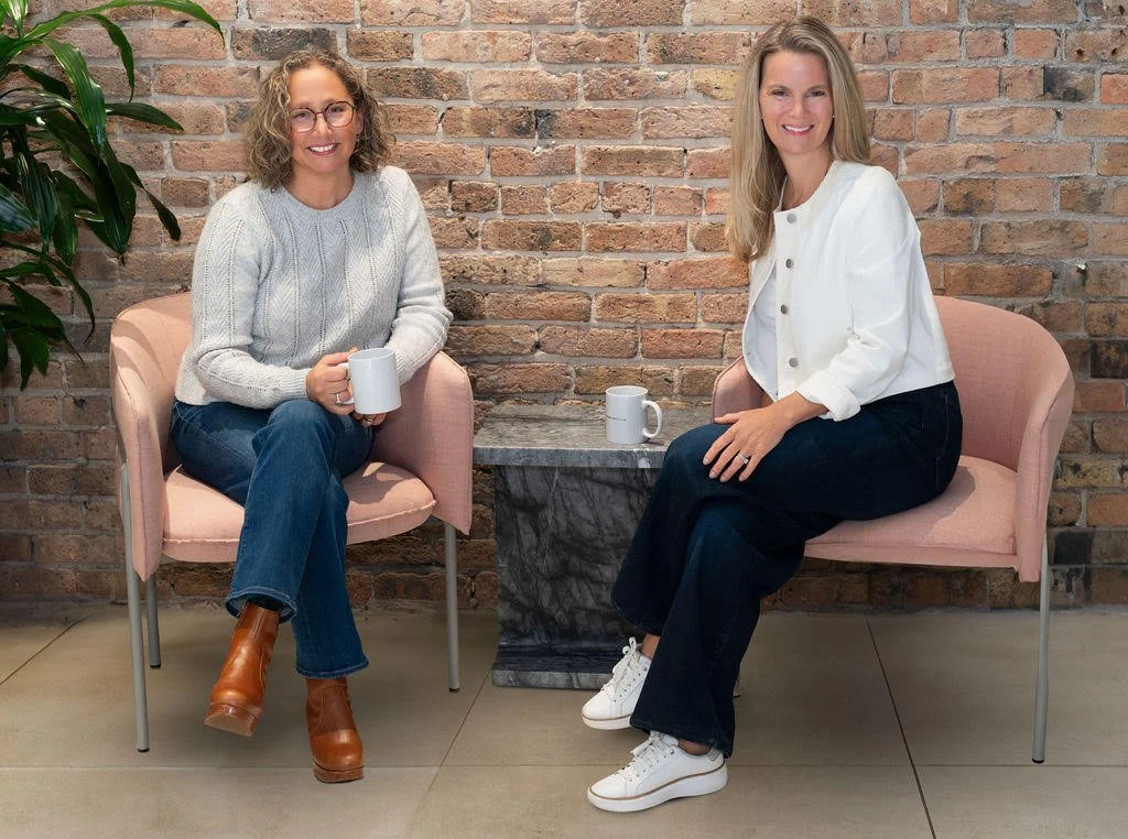 Drs. Amy and Rebecca sit in front of a red brick wall, smiling towards the camera.