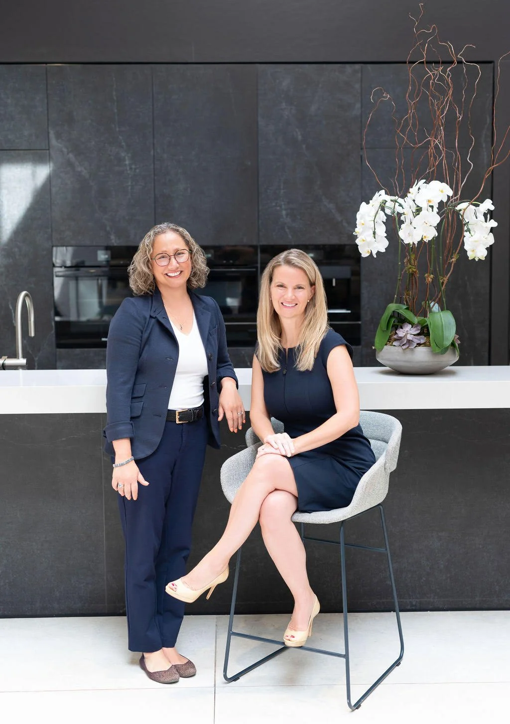 Drs. Amy and Rebecca stand in front of a modern kitchen and a bowl of orchids, smiling towards the camera.