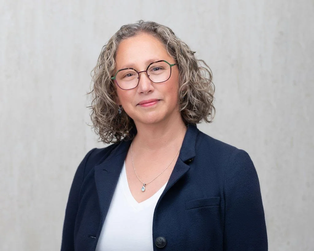 Dr. Rebecca Dunsmoor-Su is in front of a beige background, wearing a navy blazer and white shirt. She looks straight on to the camera with a slight smile.