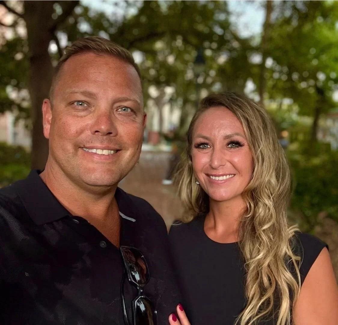 A man and a woman smiling outdoors with trees in the background.
