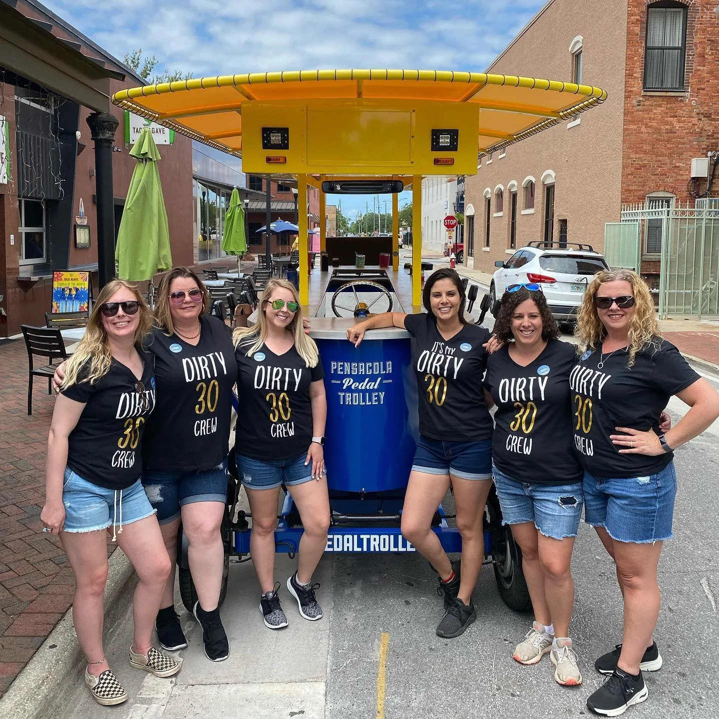 Group of six women posing in front of a pedal trolley with the sign 'Pensacola Pedal Trolley', wearing matching black shirts that read 'Dirty 30 Crew', on a city street with brick buildings and outdoor seating.