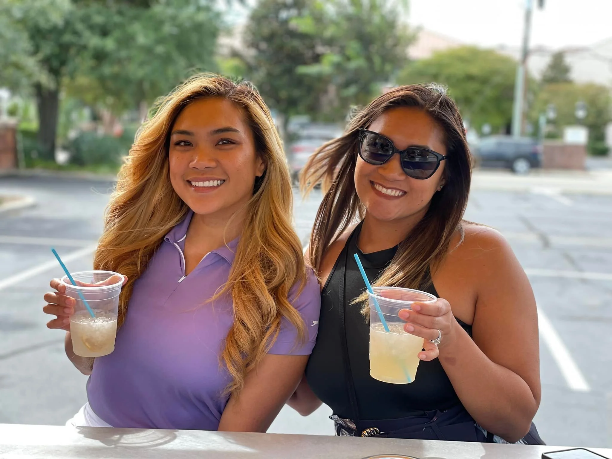 Two women smiling and holding drinks in clear cups with blue straws, standing outdoors in a parking lot with trees and cars in the background.