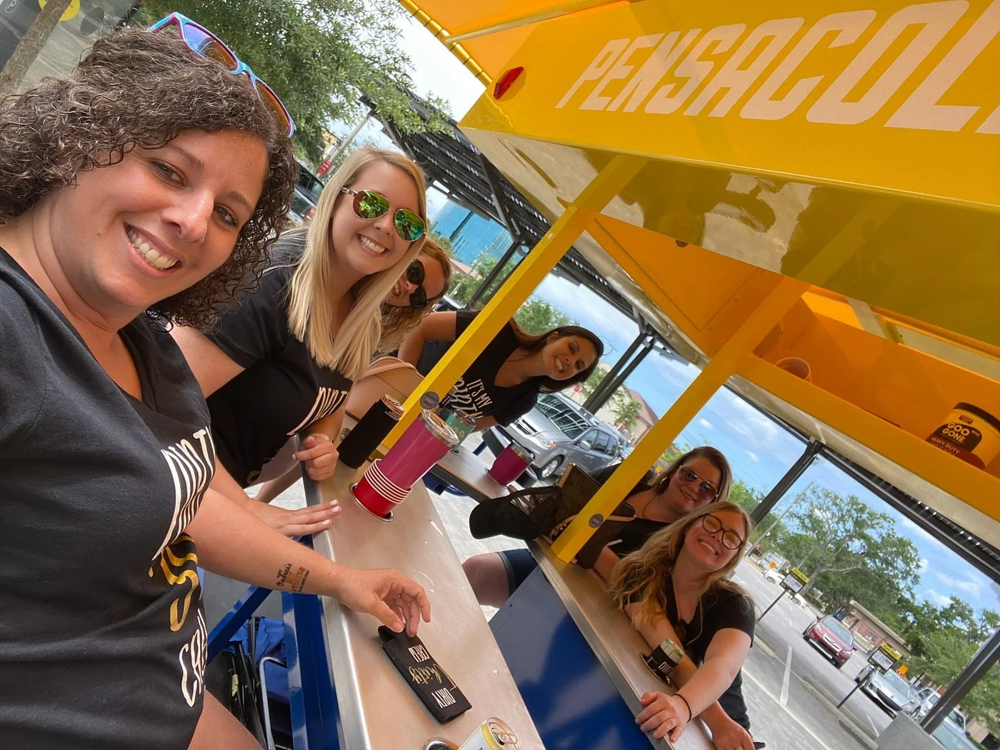 A group of six women smiling and sitting at a counter inside a yellow Pe système food truck, parked in a lot with cars and trees outside.