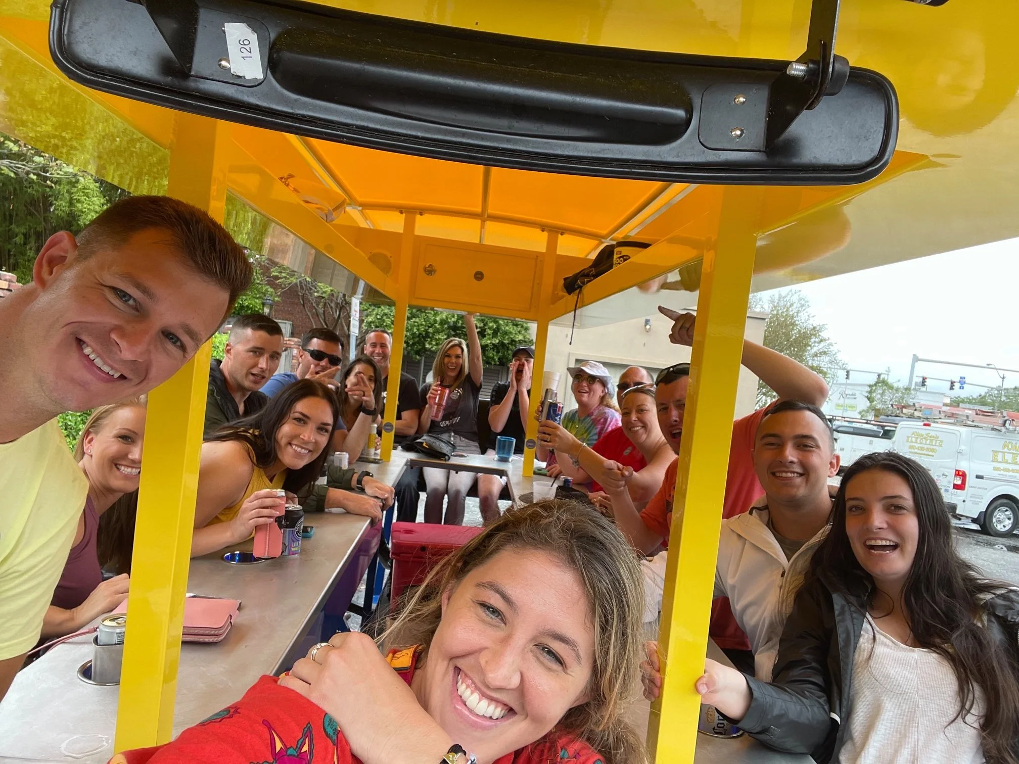 Group of people gathered under a yellow gazebo, smiling, taking selfies, and holding drinks, outdoors during the daytime.