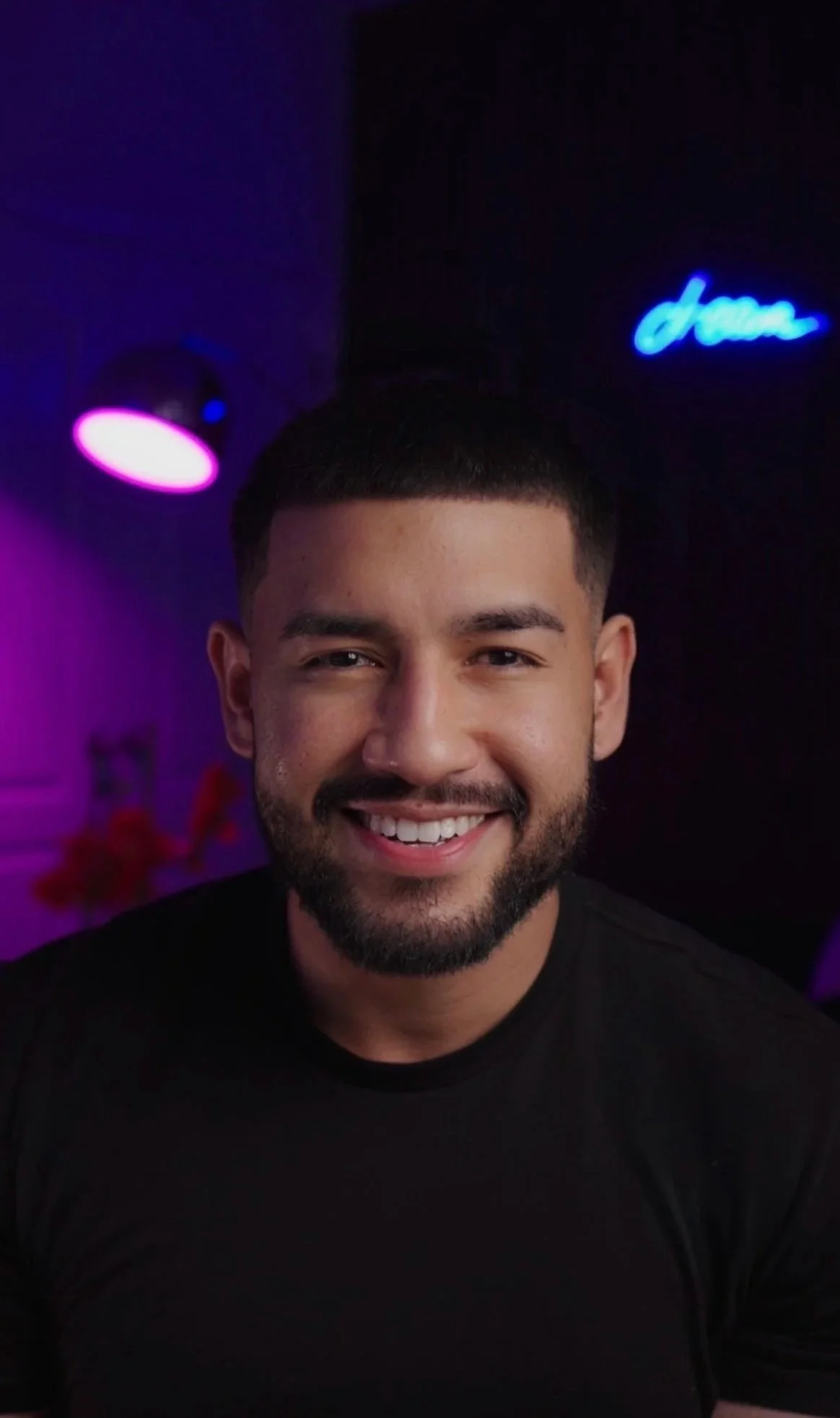 A smiling man with short dark hair, a beard, and a black shirt, illuminated by purple and pink lighting, with a neon sign in the background.