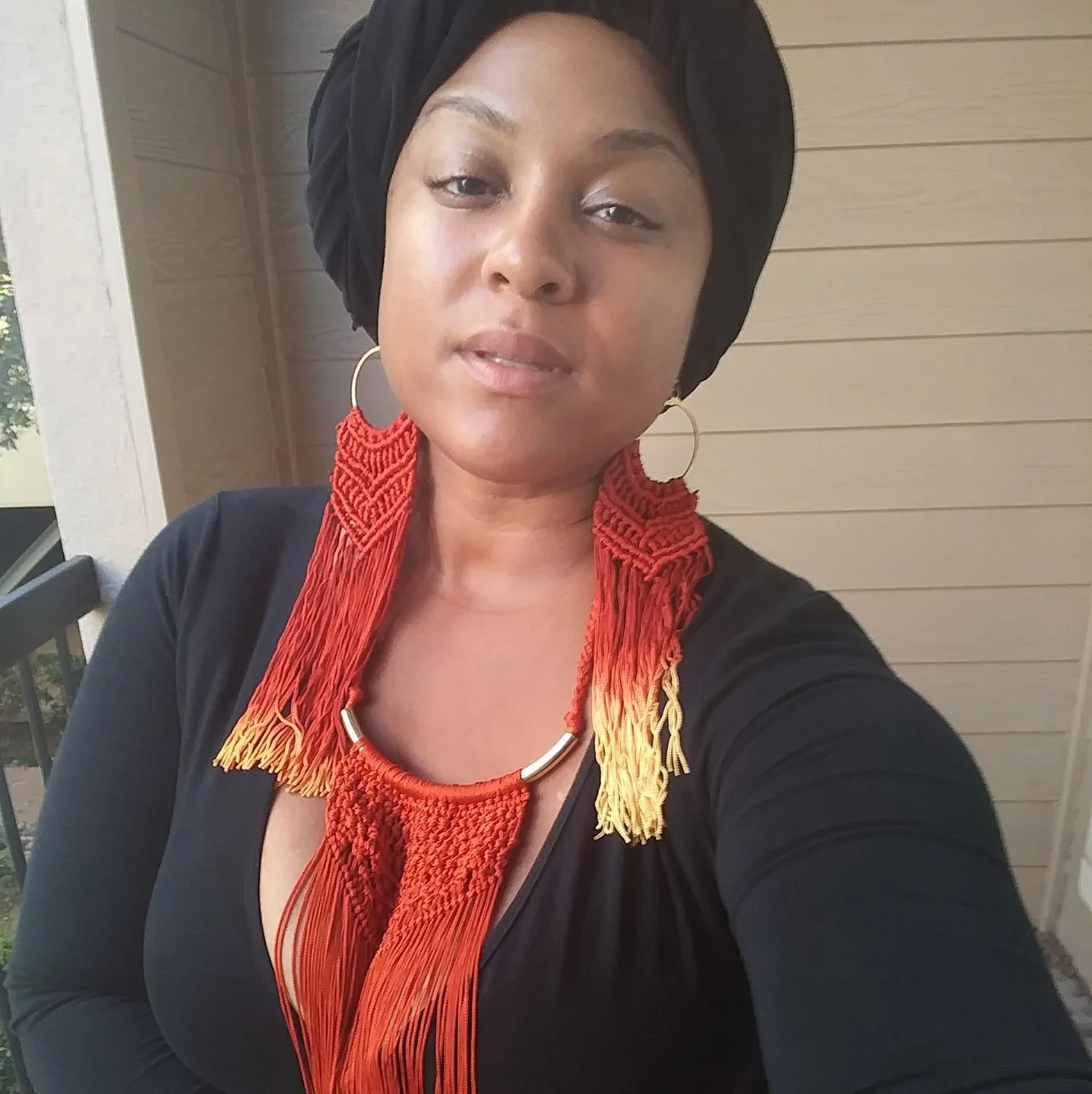A woman wearing a black head wrap, large hoop earrings, and a vibrant red, orange, and yellow woven necklace, standing outdoors in front of a beige wall with wooden siding.