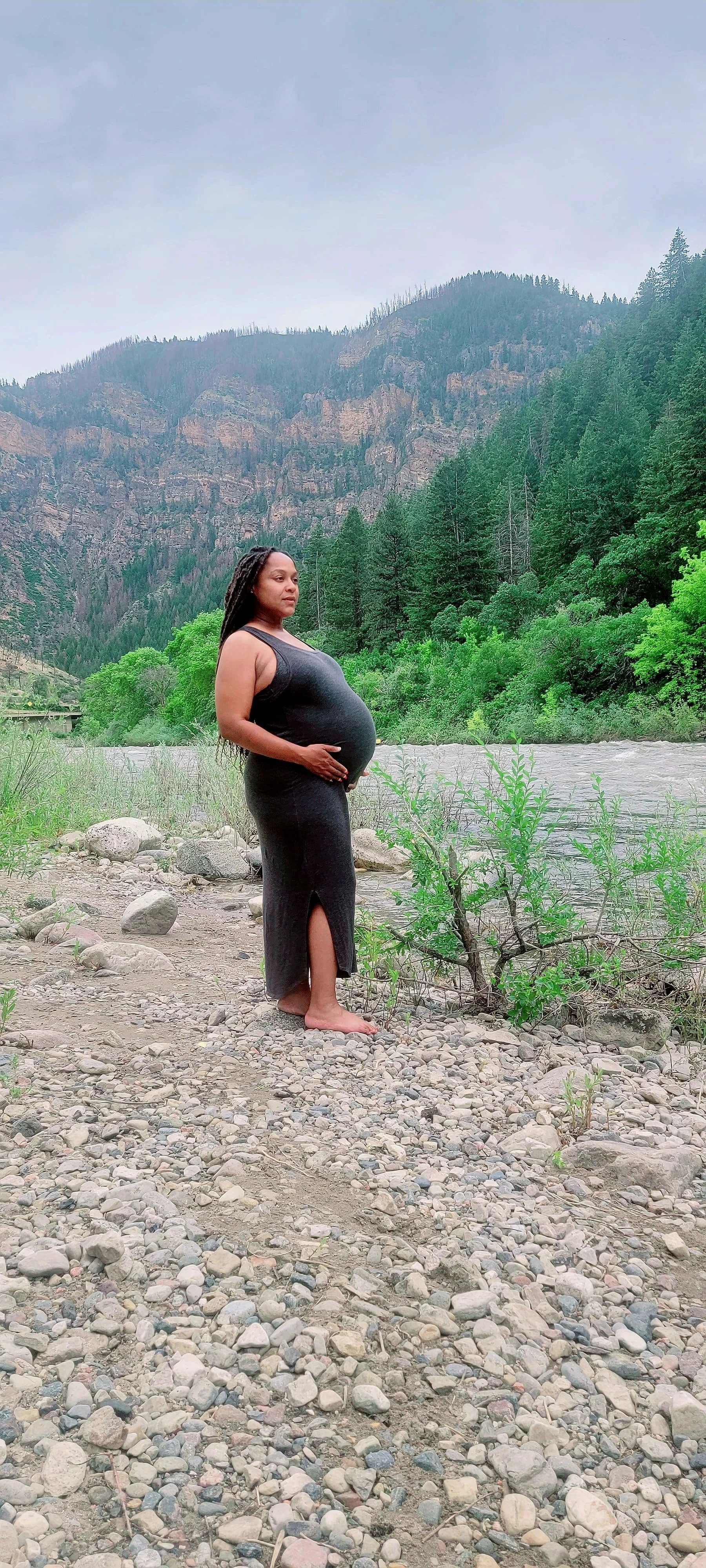 A pregnant woman in a black dress stands barefoot by a river, holding her belly, with a mountainous and forested landscape in the background.