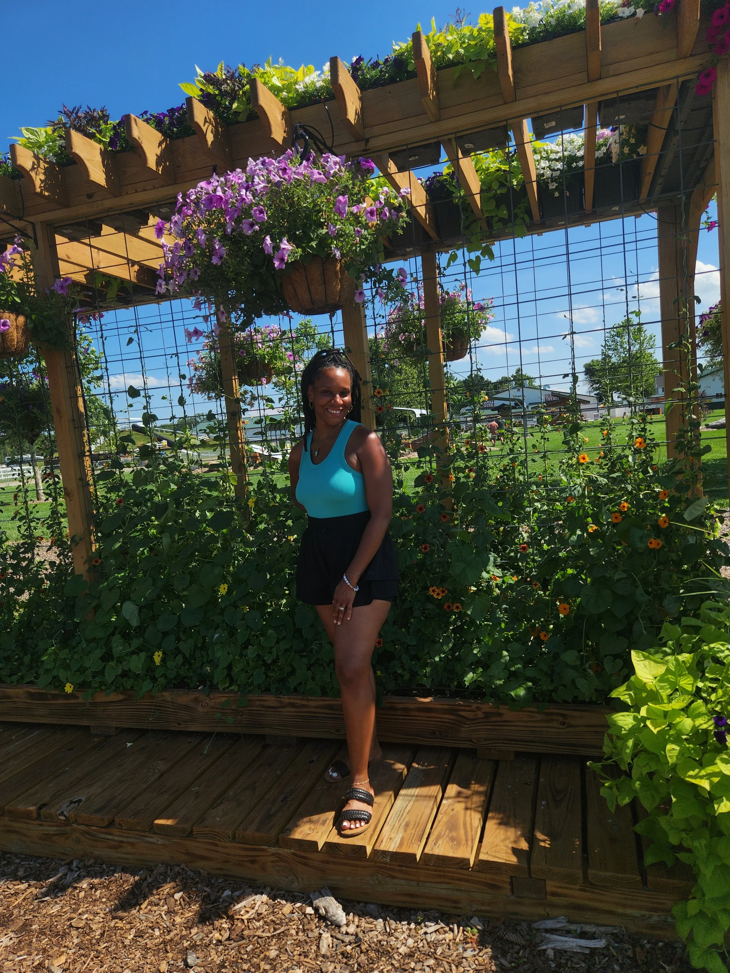 A woman standing on a wooden platform in a garden with flowering plants, a wooden trellis, and hanging baskets of purple, white, and yellow flowers under a clear blue sky.