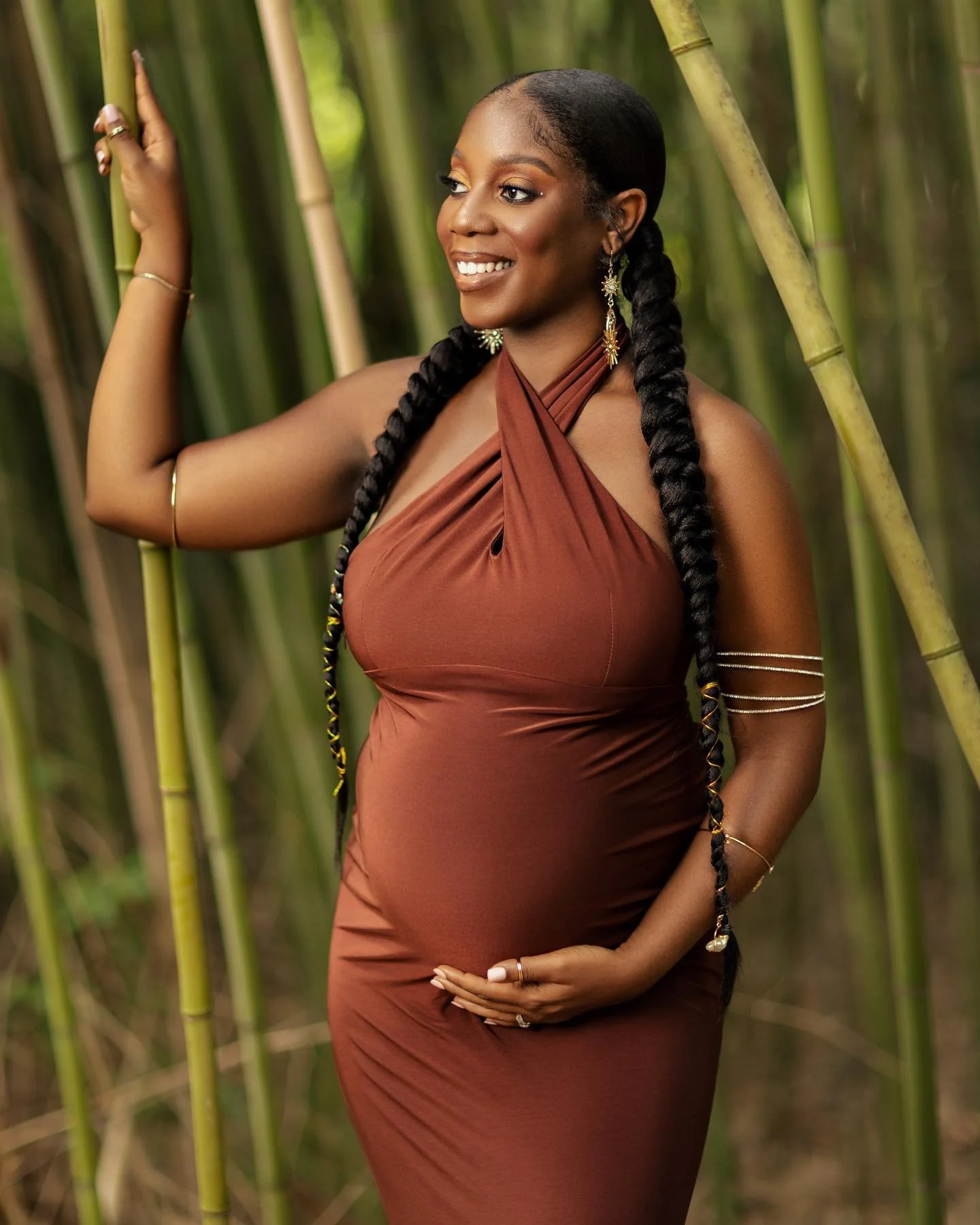 A woman wearing a brown dress with long braided hair standing among green bamboo stalks, smiling and posing with her hand raised near the bamboo.