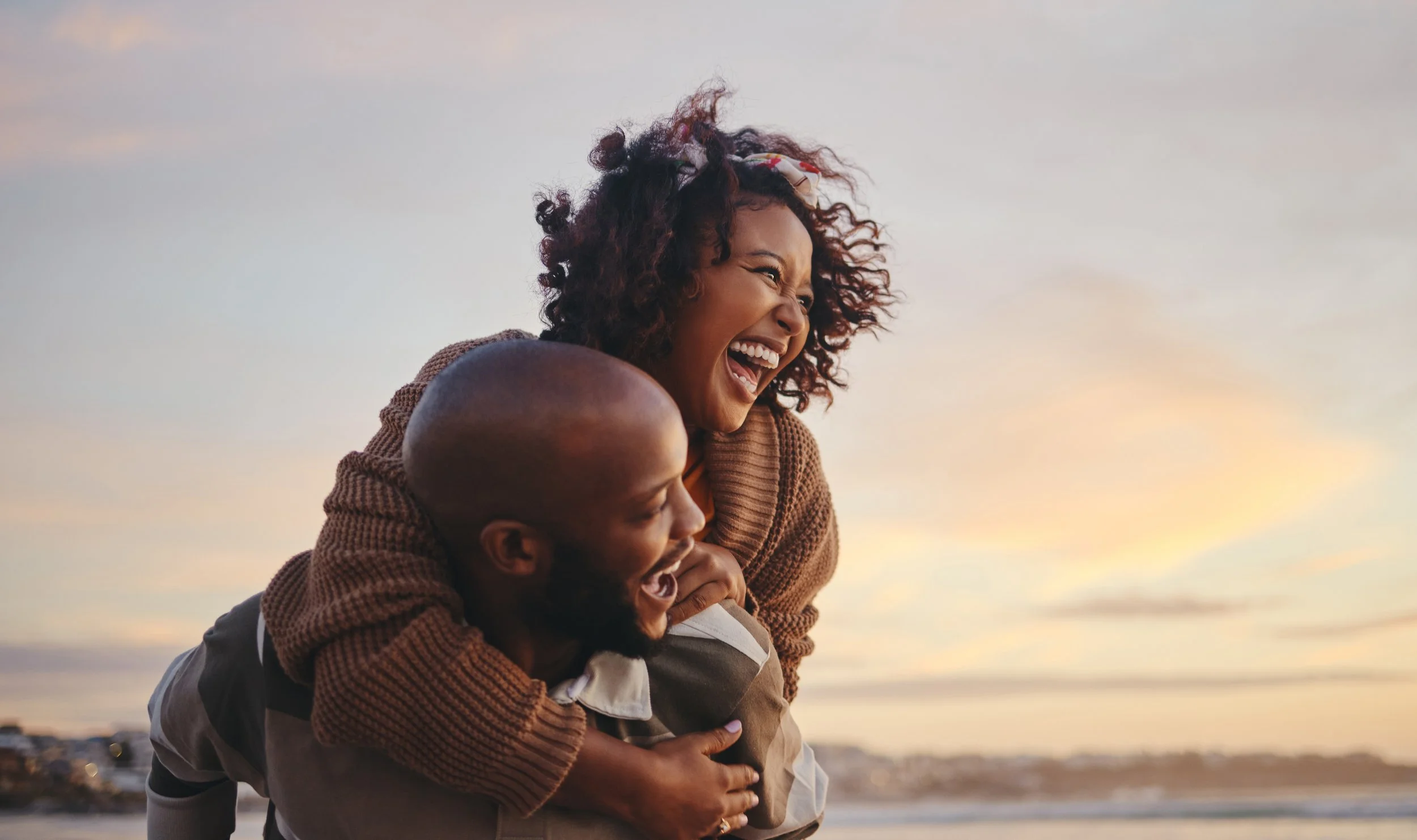 A happy woman and man enjoying a moment outdoors on the beach at sunset, with the woman playfully piggybacking on the man.