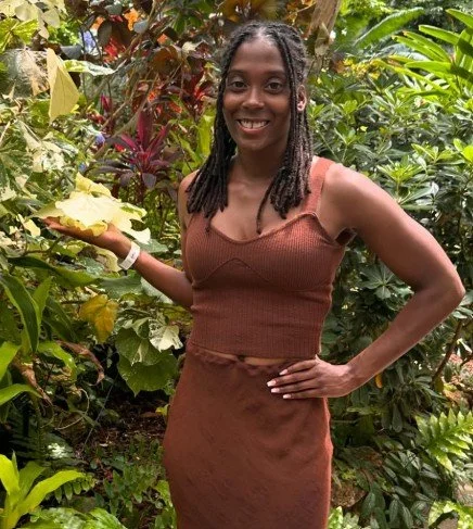 A woman standing in a lush garden surrounded by green and yellow foliage, smiling and holding some plants.