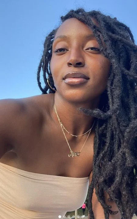 A woman with long dreadlocks taking a selfie outdoors, smiling slightly, with a clear blue sky in the background.
