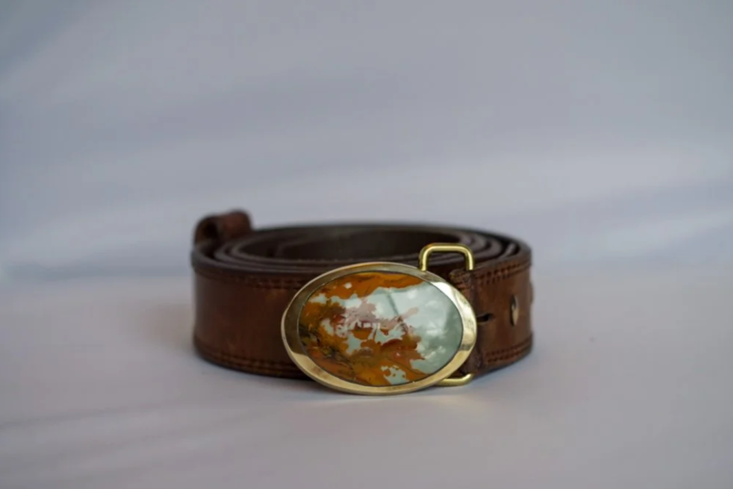 Brown leather belt with an oval gold buckle featuring a reflective inlay of a cloudy sky and autumn leaves.