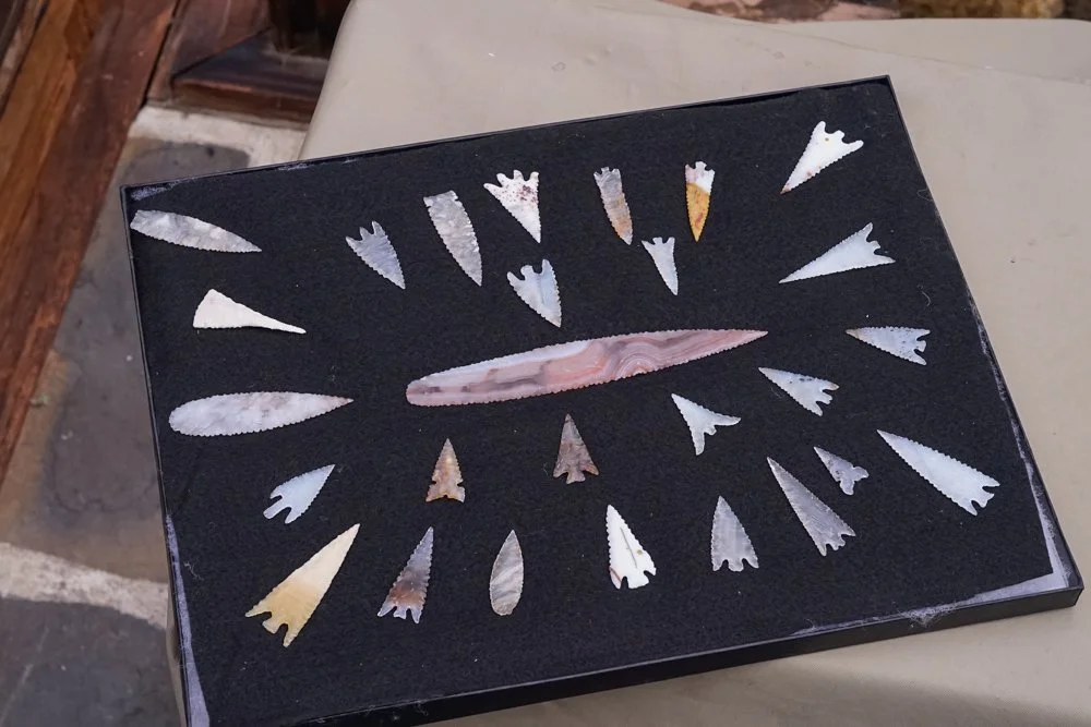 A collection of arrowhead-shaped stone tools arranged in a semi-circle on a black display box, with a central elongated stone artifact.
