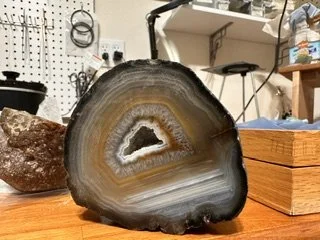 A large sliced geode with banded layers of gray, white, and brown, sitting on a wooden surface with boxes and tools in the background.