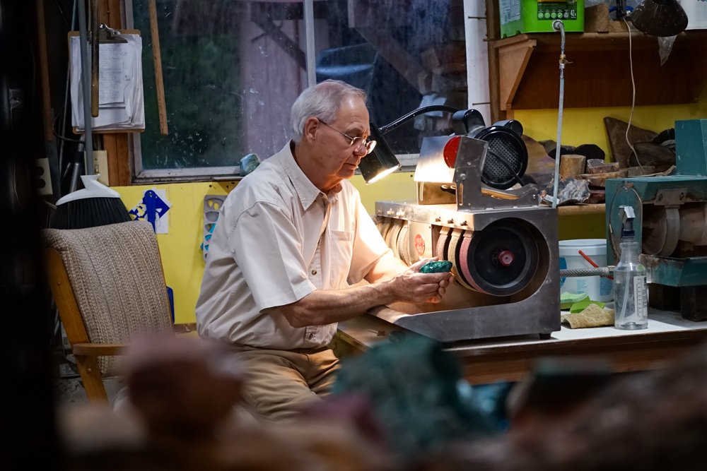 An elderly man working in a workshop, looking at his phone, with a machine on the workbench in front of him.
