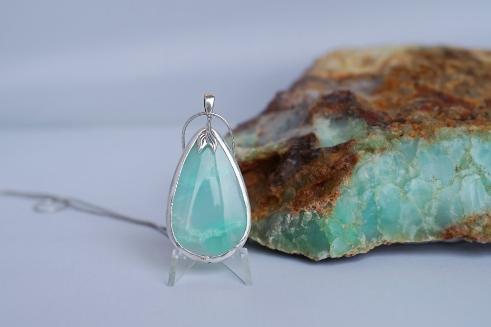 A silver pendant with a teardrop-shaped turquoise stone, placed on a small stand next to a rough turquoise rock.
