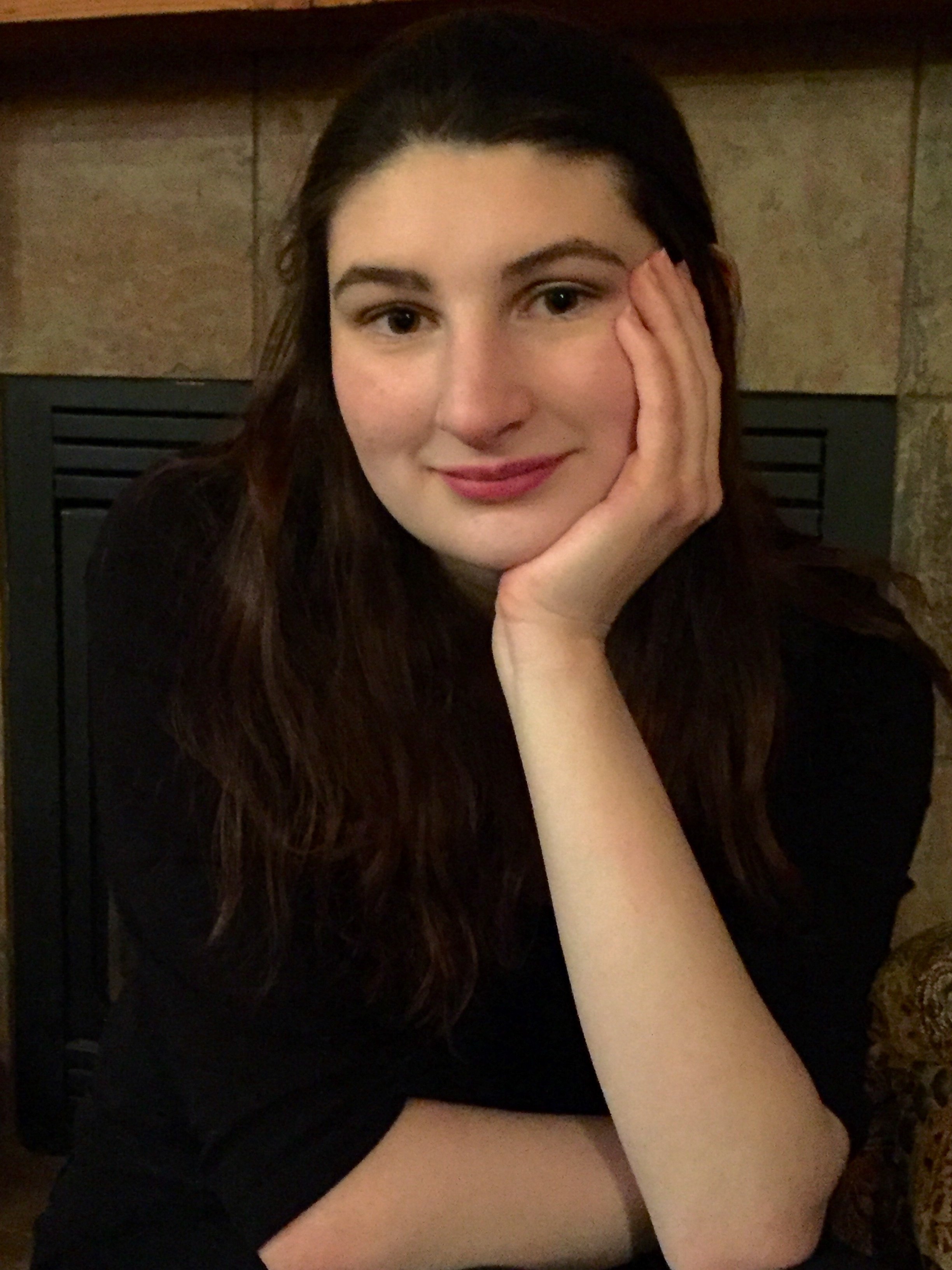 A woman with dark brown, wavy hair and light skin, resting her face on her hand, smiling softly at the camera, sitting in front of a stone fireplace.