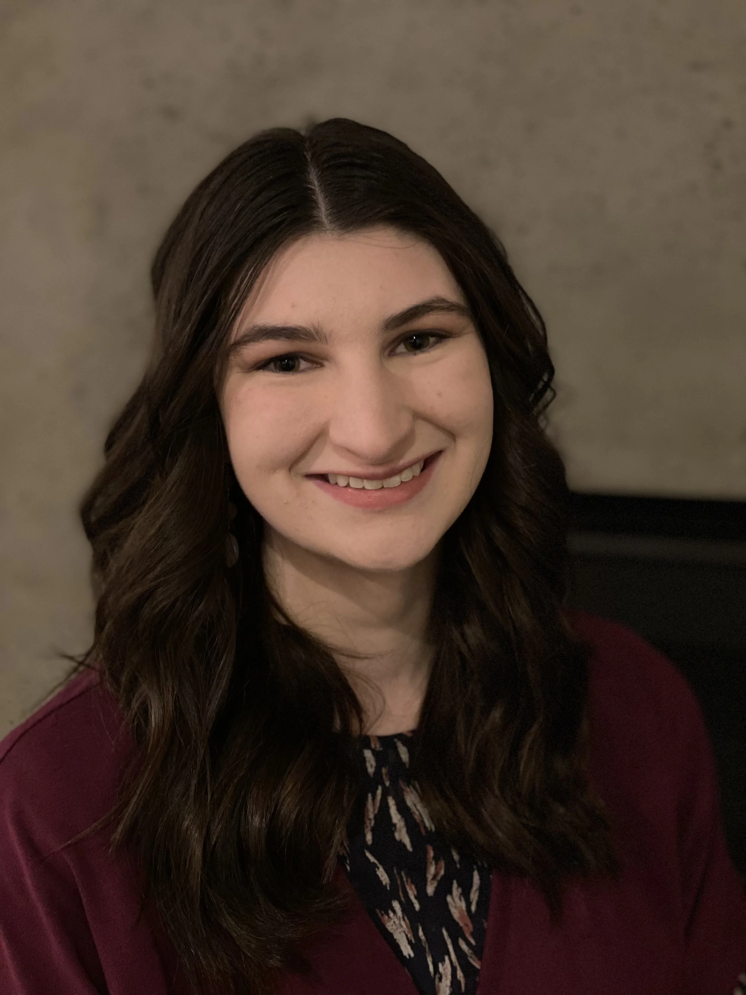 A young woman with long, wavy dark hair and light skin, smiling, wearing a maroon top with a patterned detail, against a plain background.