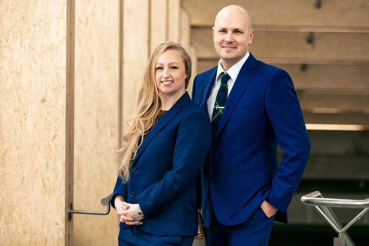 A woman and a man in blue suits standing in front of a wooden background, smiling for the camera.