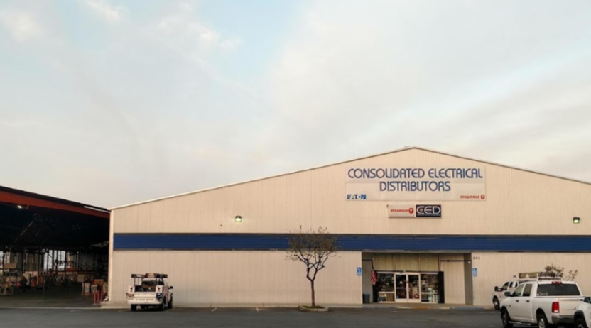 Large warehouse building with sign 'Consolidated Electrical Distributors.' Parking lot in front with three vehicles parked near the entrance. A small tree environment in front of the building.