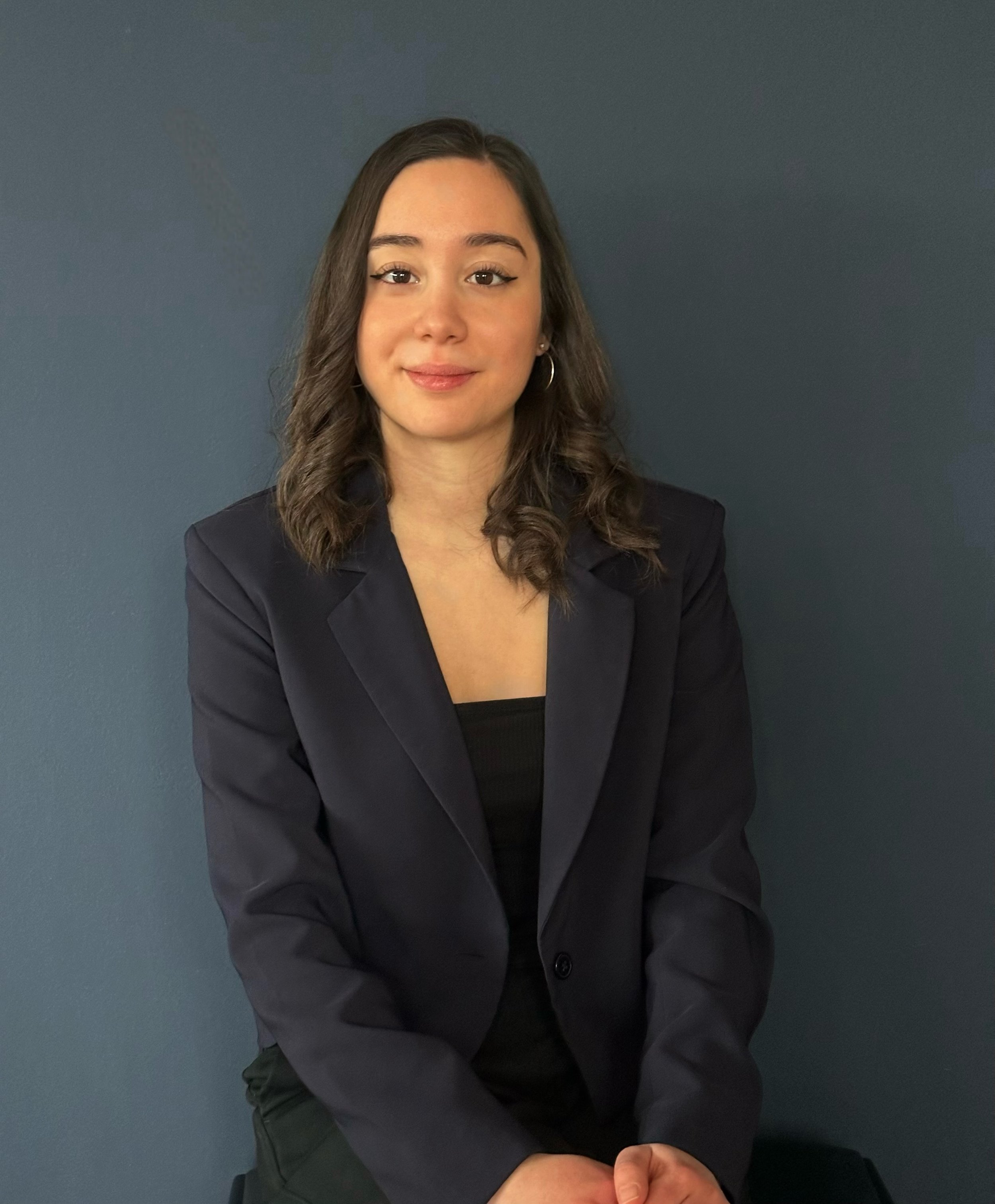 Junge Frau mit braunen Haaren in dunkler Blazer sitzt vor einer blauen Wand.