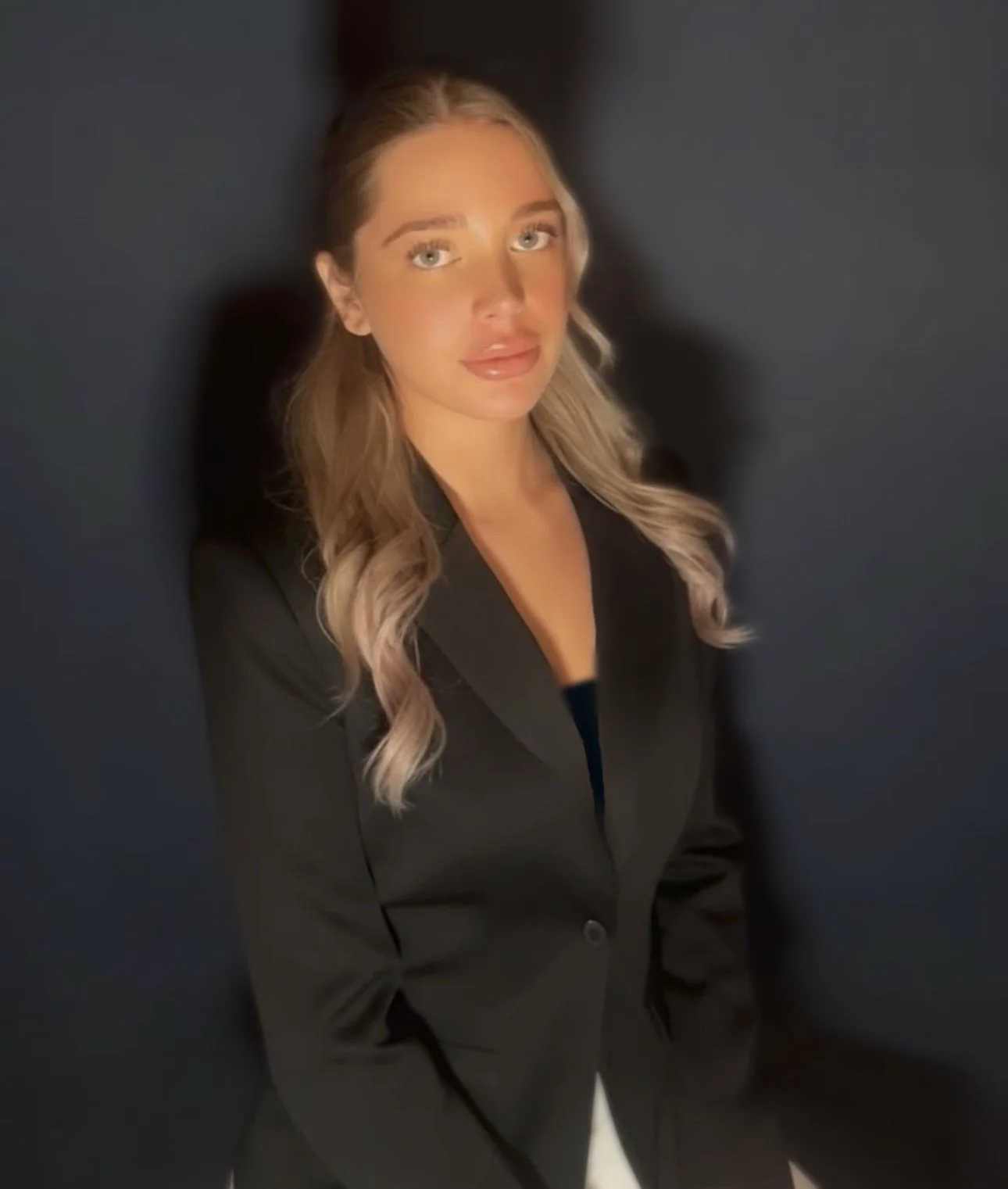 Junge Frau mit langen blonden Haaren im schwarzen Blazer vor dunklem Hintergrund.